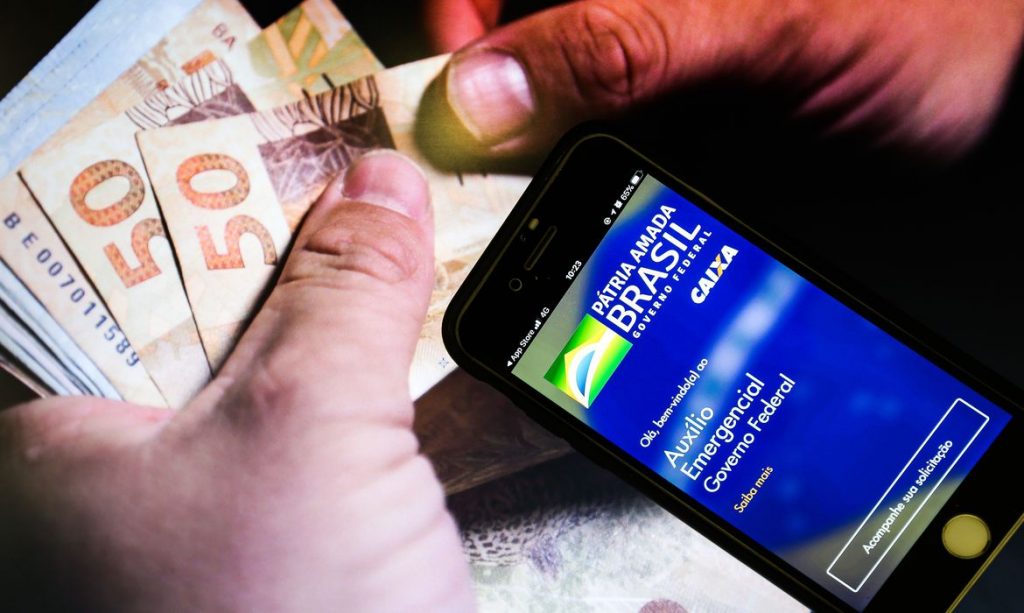 Dinheiro sendo contado, ao lado, um celular com o aplicativo Auxílio Emergencial do Governo Federal
